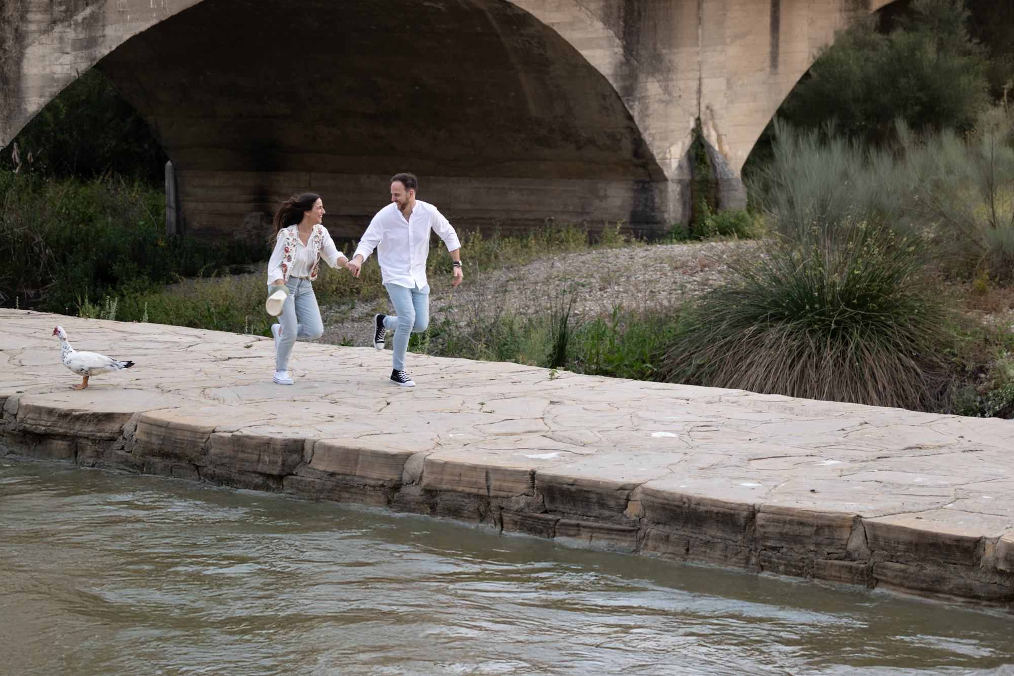 Novios corriendo junto a un río y un puente de fonod. Sesión Pre Boda