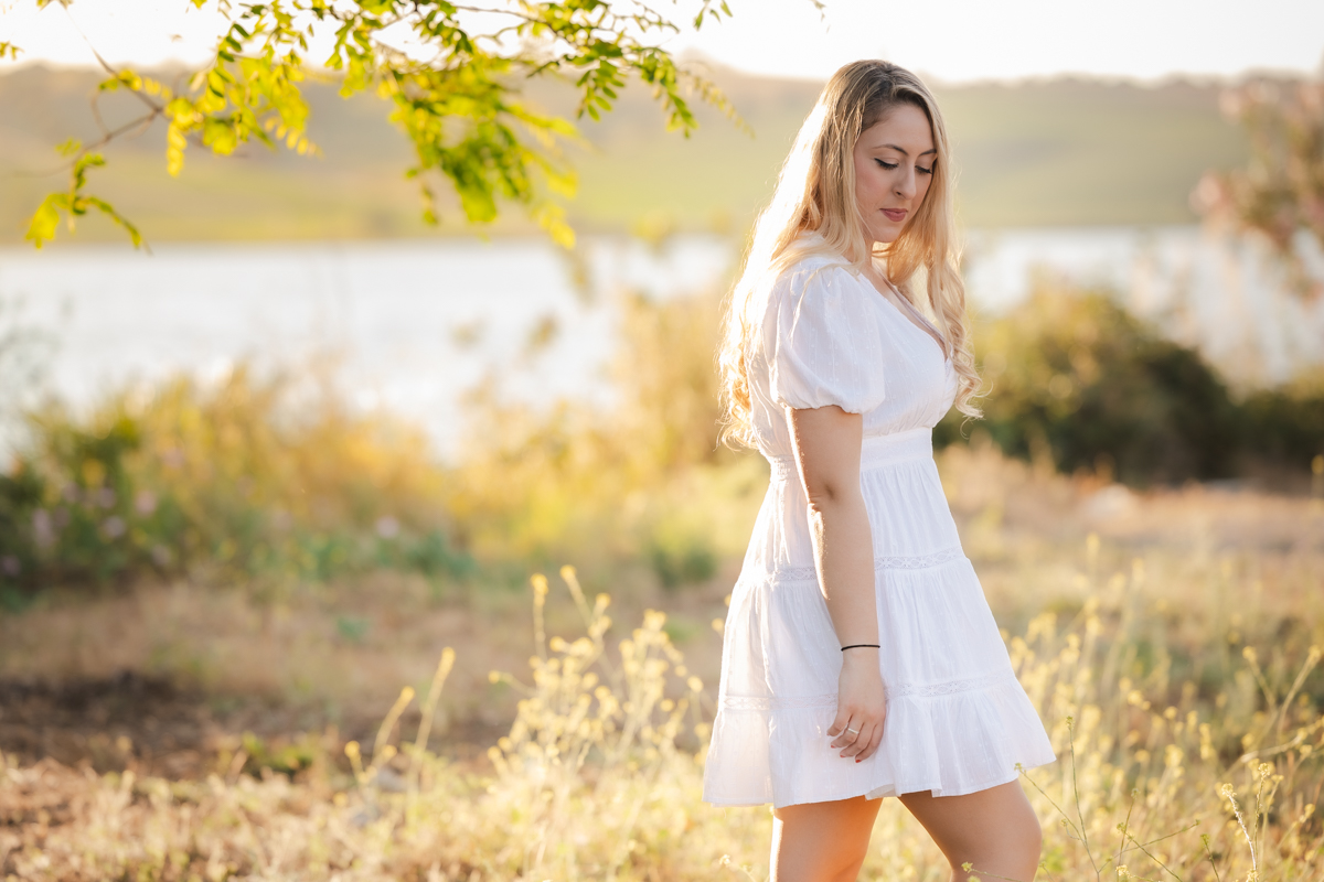 Mujer de pelo rubio y vestida de blanco junto a un lago al atardecer