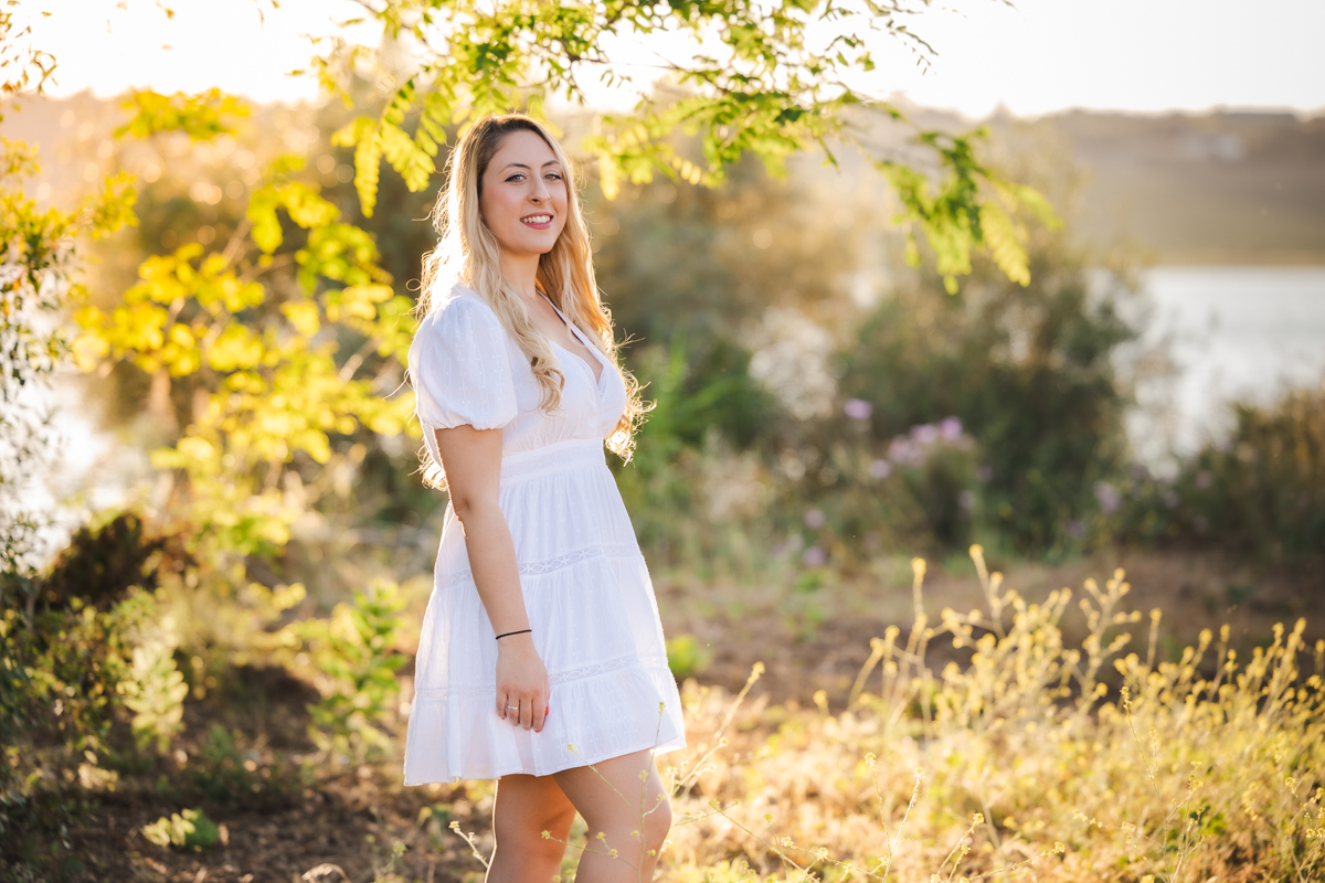 Mujer de pelo rubio y vestida de blanco junto a un lago al atardecer