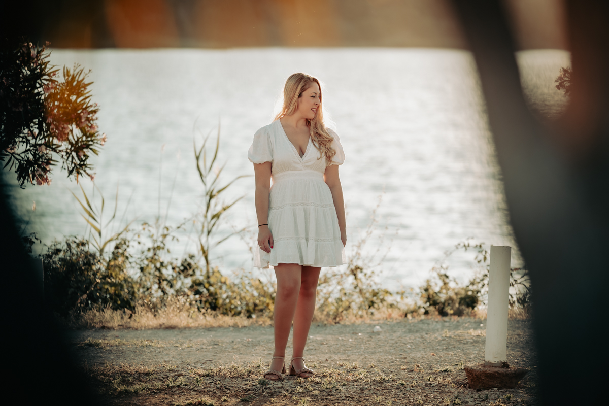 Mujer de mediana edad, de pelo rubio, vestido blanco, posa de cuerpo entero junto al lago de Arcos