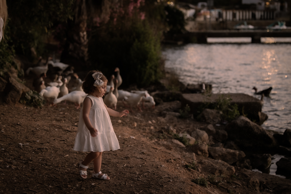 Niña pequeña vestida de blanco juega con unos patos junto al lago de Arcos